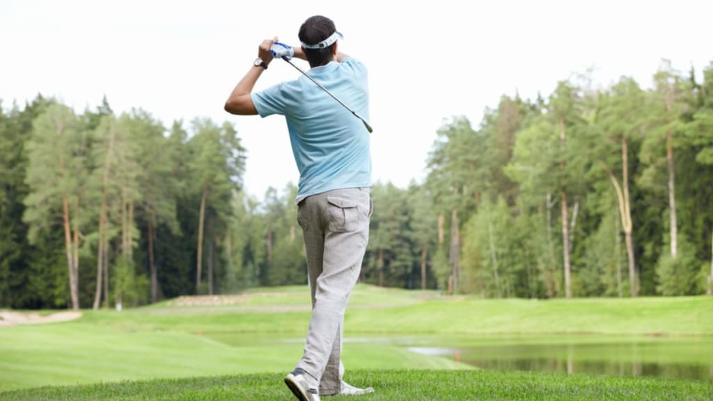 A person wearing a blue shirt and gray pants is captured from behind, swinging a golf club on a lush green golf course surrounded by trees.